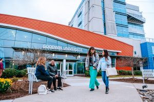 Two students walk by Goodwin University's main campus on One Riverside Drive, while two other students laugh on a bench near the building's entrance.