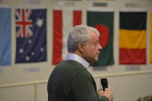 Goodwin University President and Founder Mark E. Scheinberg speaks into a microphone, photographed in profile with a series of international flags in the background.