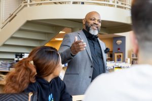 A man in a gray blazer, black top, and black bow tie smiles while engaging with a table of students at Goodwin University's Hoffman Family Library.