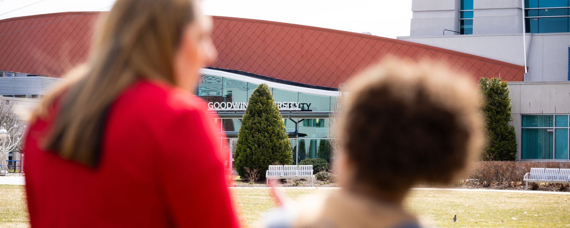 Two women stand outside Goodwin University, seen from behind and slightly out of focus, while the Goodwin University building sign comes into clear view in front of them.