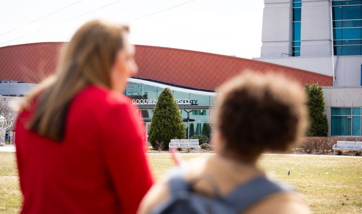 Two women stand outside Goodwin University, seen from behind and slightly out of focus, while the Goodwin University building sign comes into clear view in front of them.