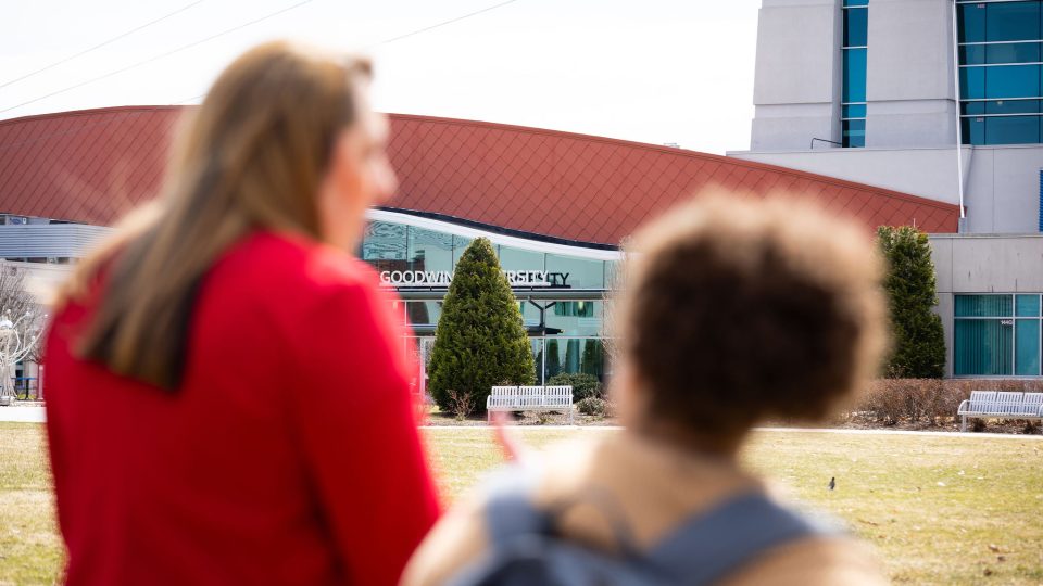 Two women stand outside Goodwin University, seen from behind and slightly out of focus, while the Goodwin University building sign comes into clear view in front of them.