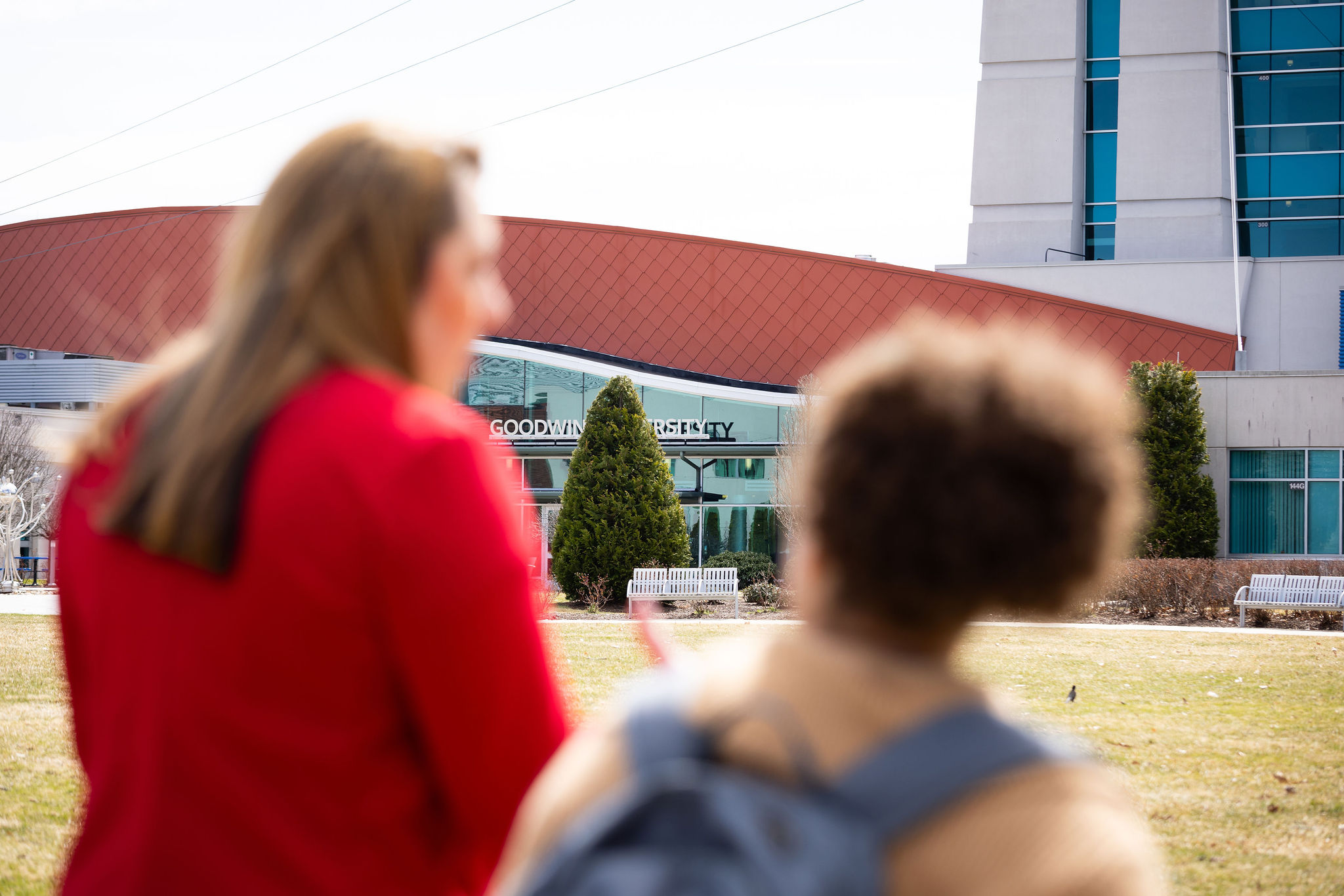 Two women stand outside Goodwin University, seen from behind and slightly out of focus, while the Goodwin University building sign comes into clear view in front of them.