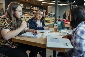 Three students sit together at a table in a coffee shop with textbooks open.