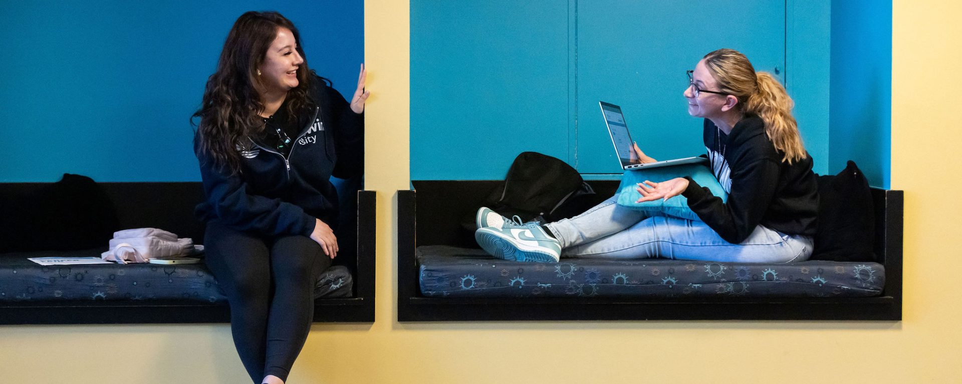 Two students sit in blue seating nooks, talking as they work.