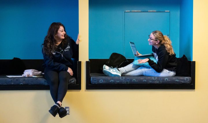 Two students sit in blue seating nooks, talking as they work.