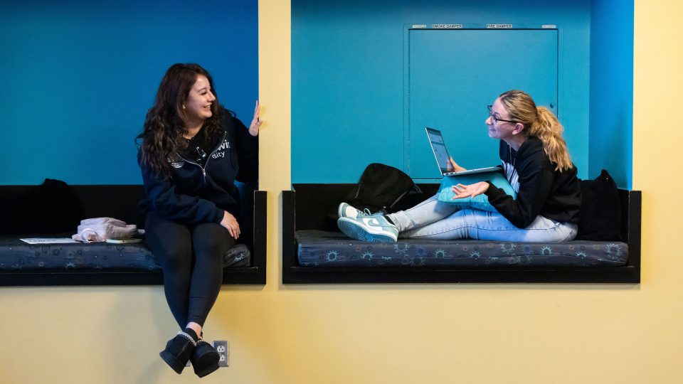 Two students sit in blue seating nooks, talking as they work.