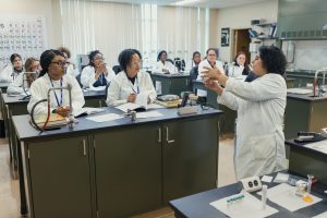 Students in white lab coats watch their professor give a lecture. 