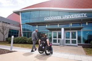 A man walks alongside a woman in a wheelchair toward Goodwin University’s main entrance.
