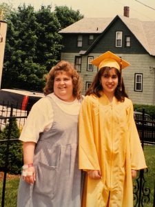 A mother smiles next to her daughter, who is dressed in yellow graduation regalia.