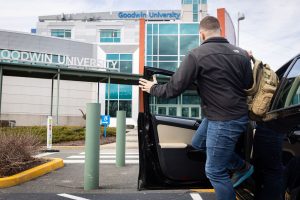 A student with a backpack over their shoulder exits a car in front of the main entrance to Goodwin University.