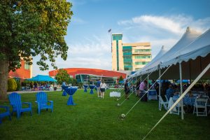 Outdoor gathering beside a large, white tent on a sunny day, with blue tables and chairs on green grass, near a modern building with a tall tower.