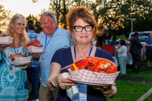 A woman holds a large, cooked lobster on a plate at an outdoor gathering
