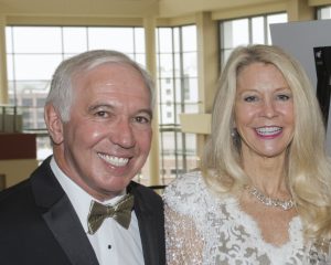 A smiling man in a tuxedo and bow tie sits beside a woman in a white lace dress and necklace
