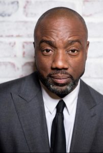 A man in a gray suit and black tie stands against a white brick wall