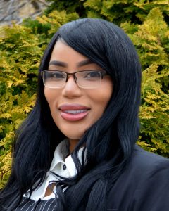 A woman with long black hair, glasses, and a black blazer is standing outdoors in front of green bushes, smiling at the camera.