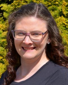 A woman with wavy brown hair, glasses, and a black t-shirt is standing outdoors in front of green bushes, smiling at the camera.