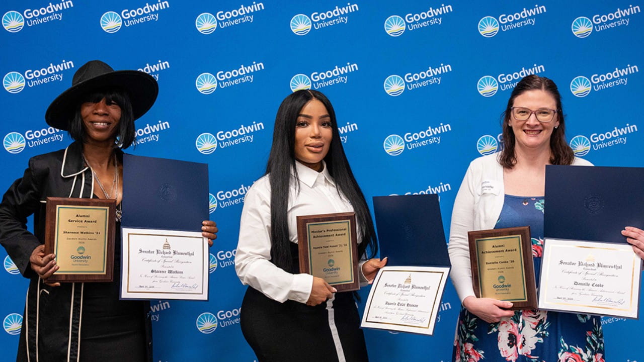 Three women stand in front of a blue Goodwin University backdrop, each holding plaques and certificates, posing for a photo at an award ceremony.