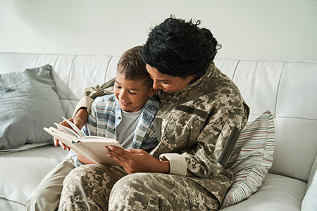 A female veteran and a young boy read a book together while sitting on a couch.